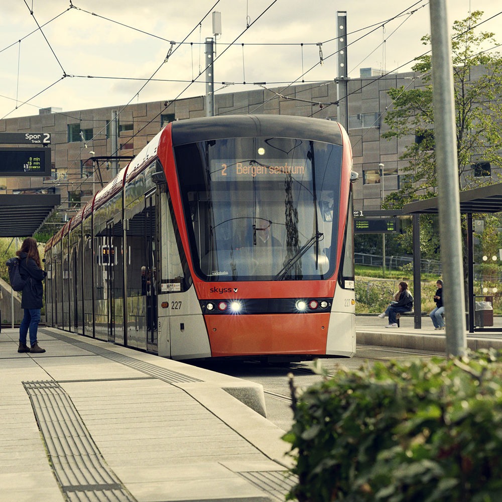 Bildet viser ei bybanevogn som står i ro på ein haldeplass, med folk som ventar på plattformen for å gå om bord.