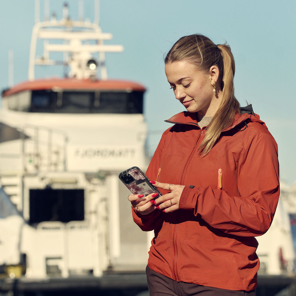 Ung kvinne ser på telefonen der ho står i fint vêr, på ei kai med ein passasjerbåt i bakgrunnen.